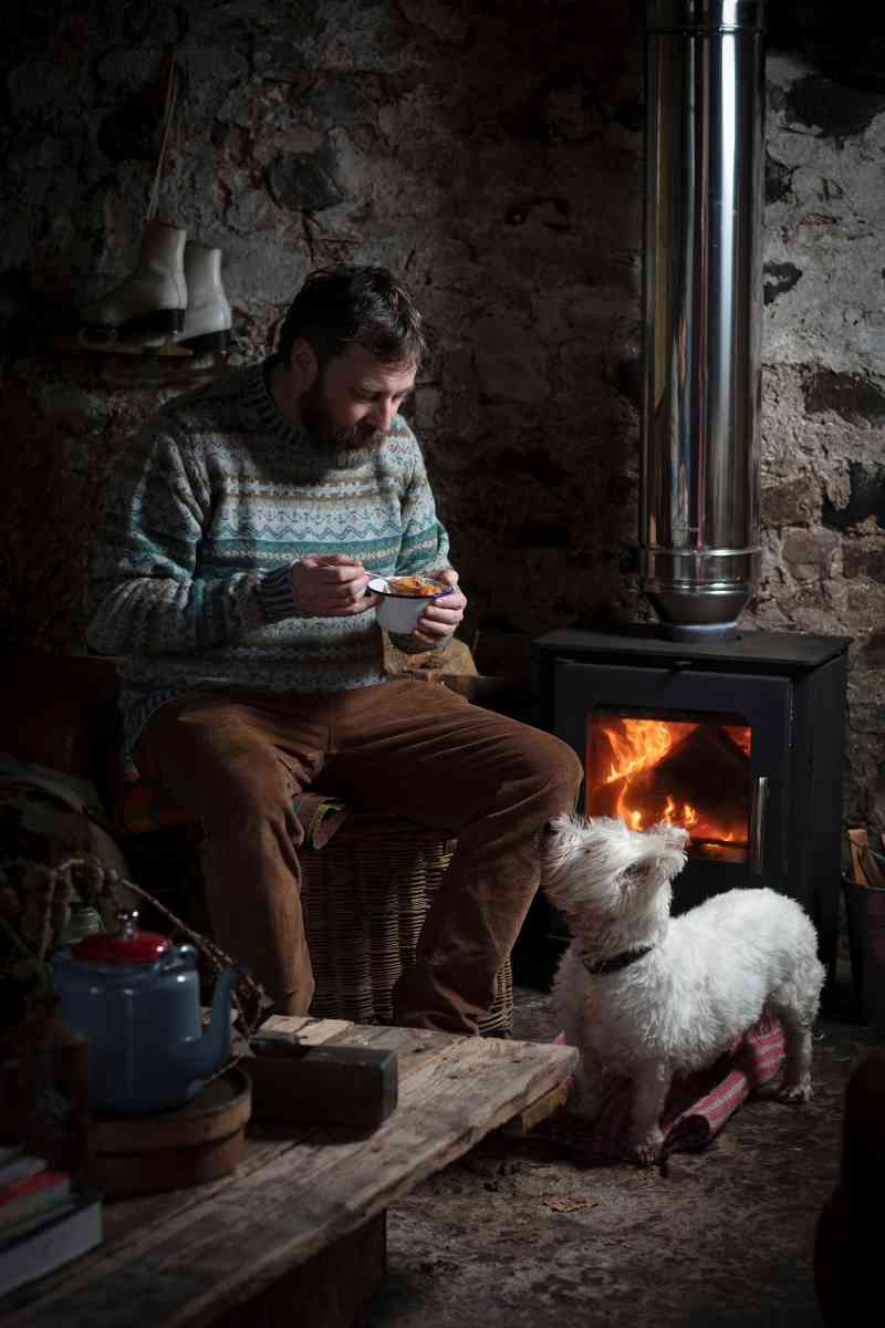 Peter MacQueen disfruta de un caldo escocés junto al fuego con su West Highland terrier, Seòras, en Guardswell Farm en Perthshire. MacQueen es el catador oficial de recetas de su compañero Coinneach MacLeod y la foto fue tomada durante una sesión de fotos para su nuevo libro